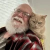 A smiling man with a white beard and glasses has a light-colored cat perched on his shoulder.