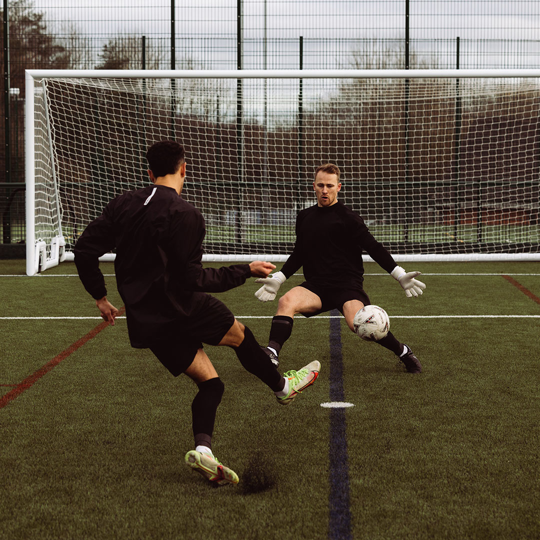 Soccer player kicking ball towards goalkeeper on a field with goal net in background.