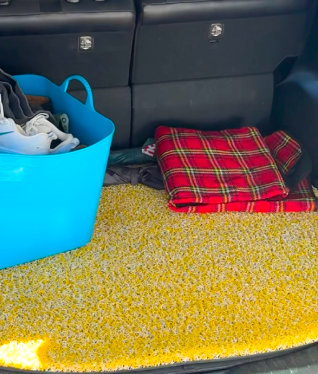 The trunk of a car with a yellow floor mat, a blue tub, and a red plaid blanket.