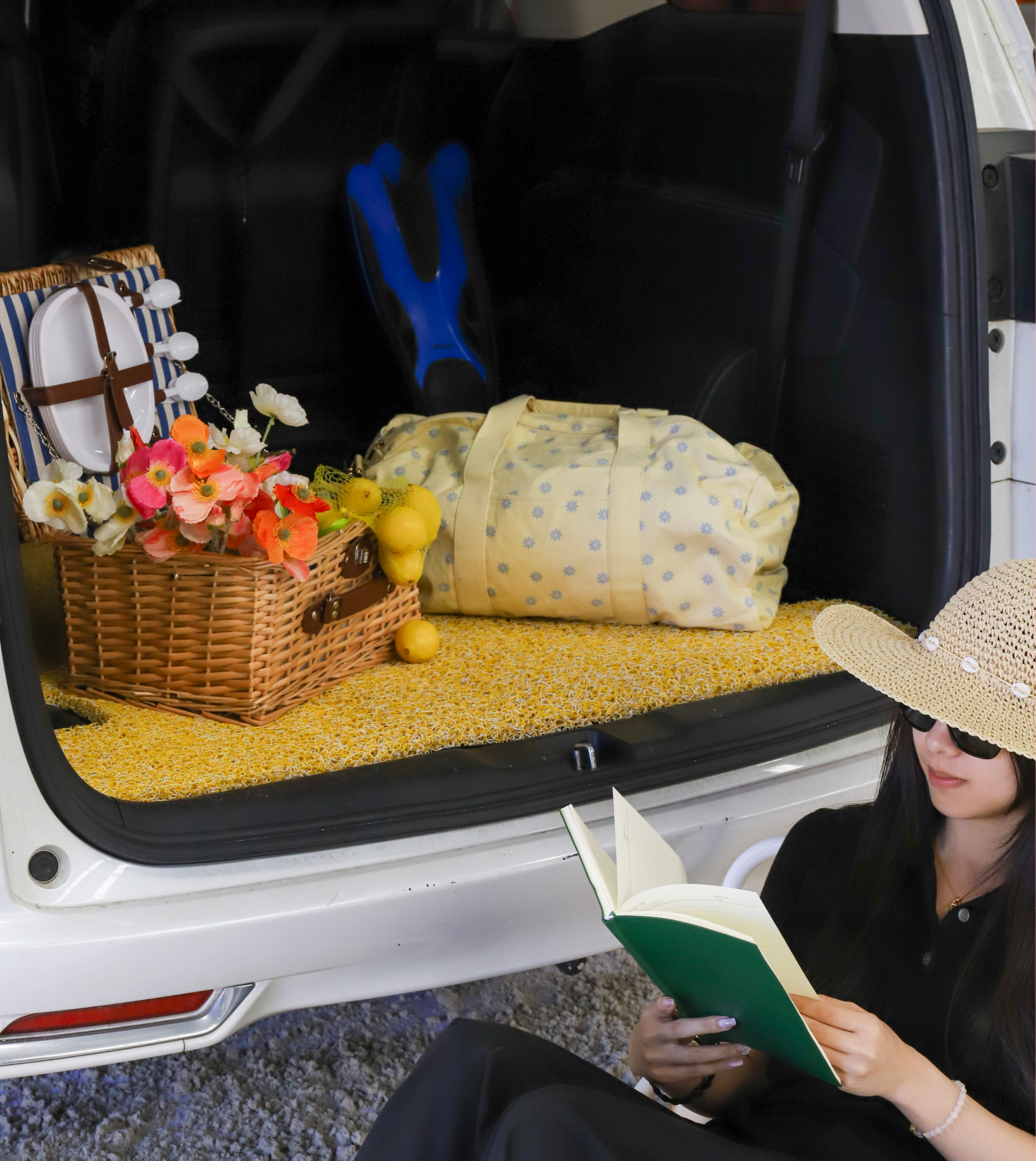 Person reading a book next to an open car trunk with a picnic basket and bags.
