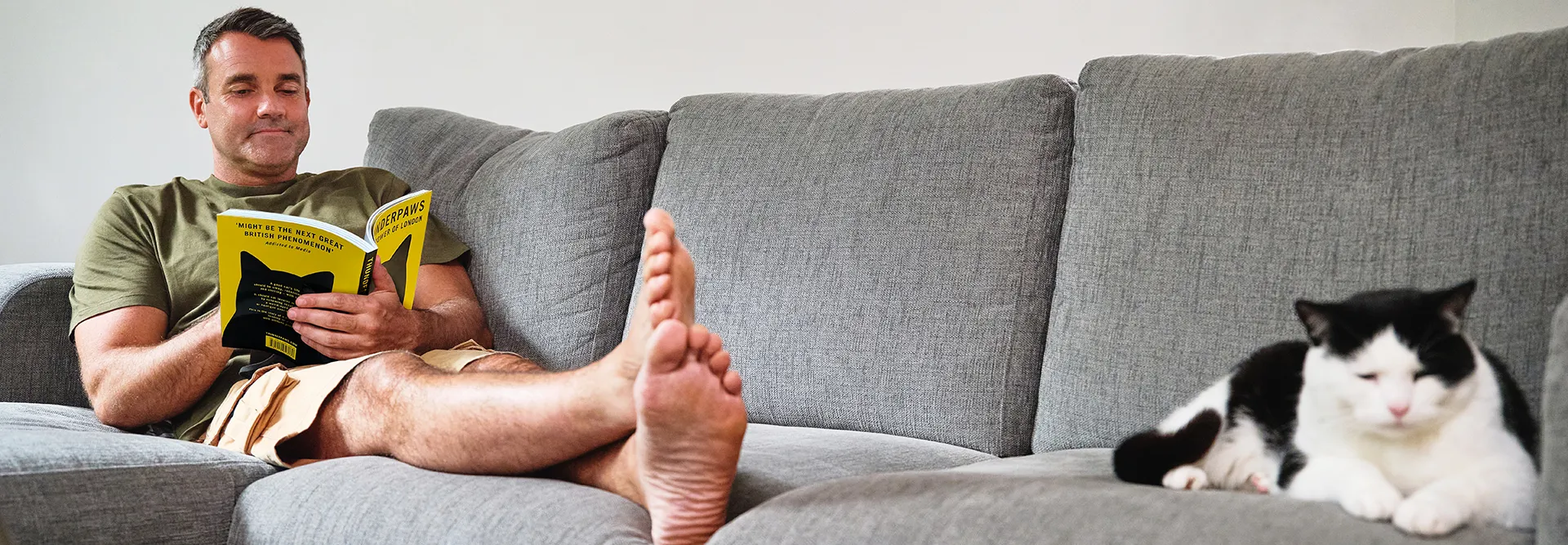 Man reading a book on a gray sofa with a black and white cat.