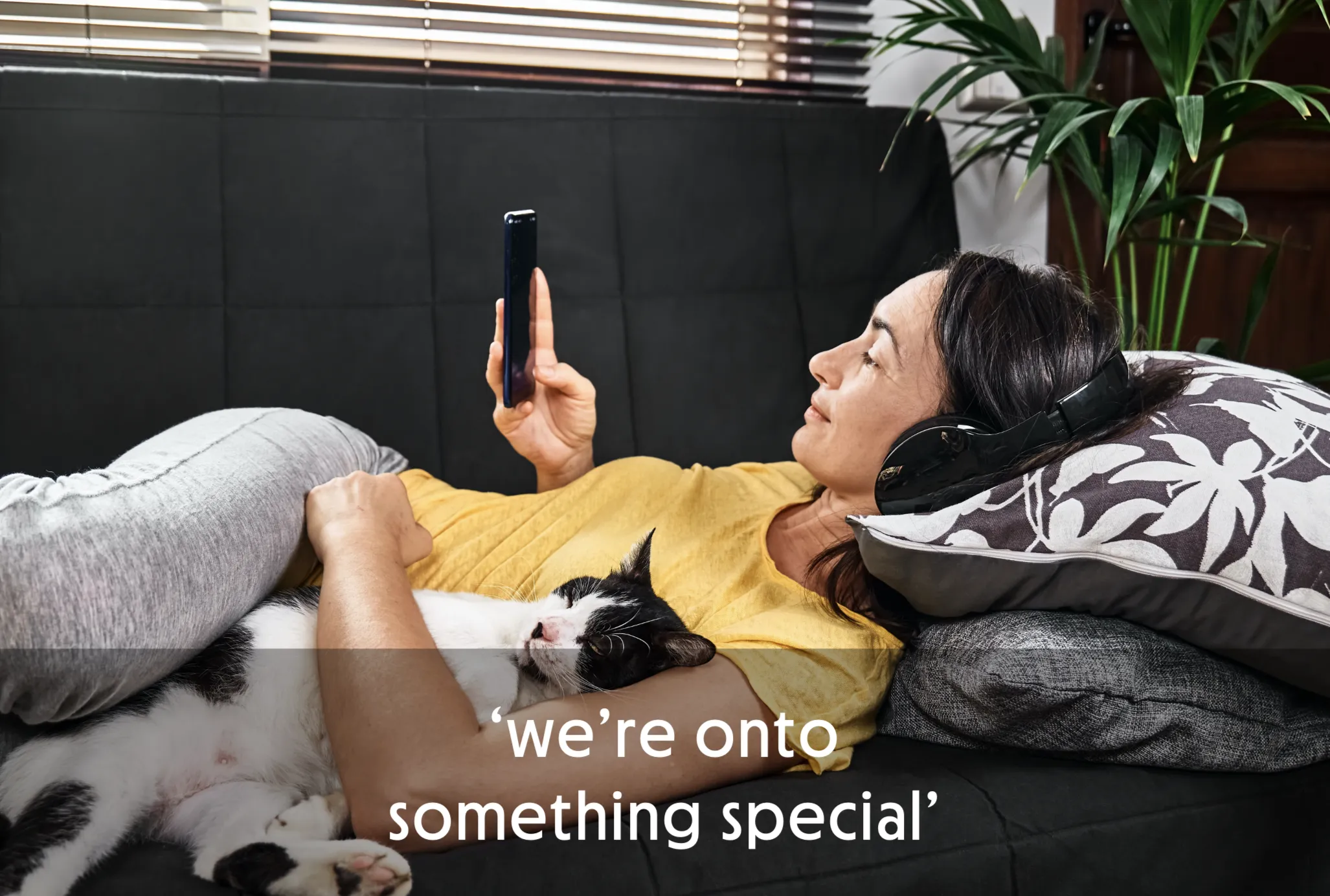 Woman lying on couch with headphones, holding phone, cat resting on her chest.