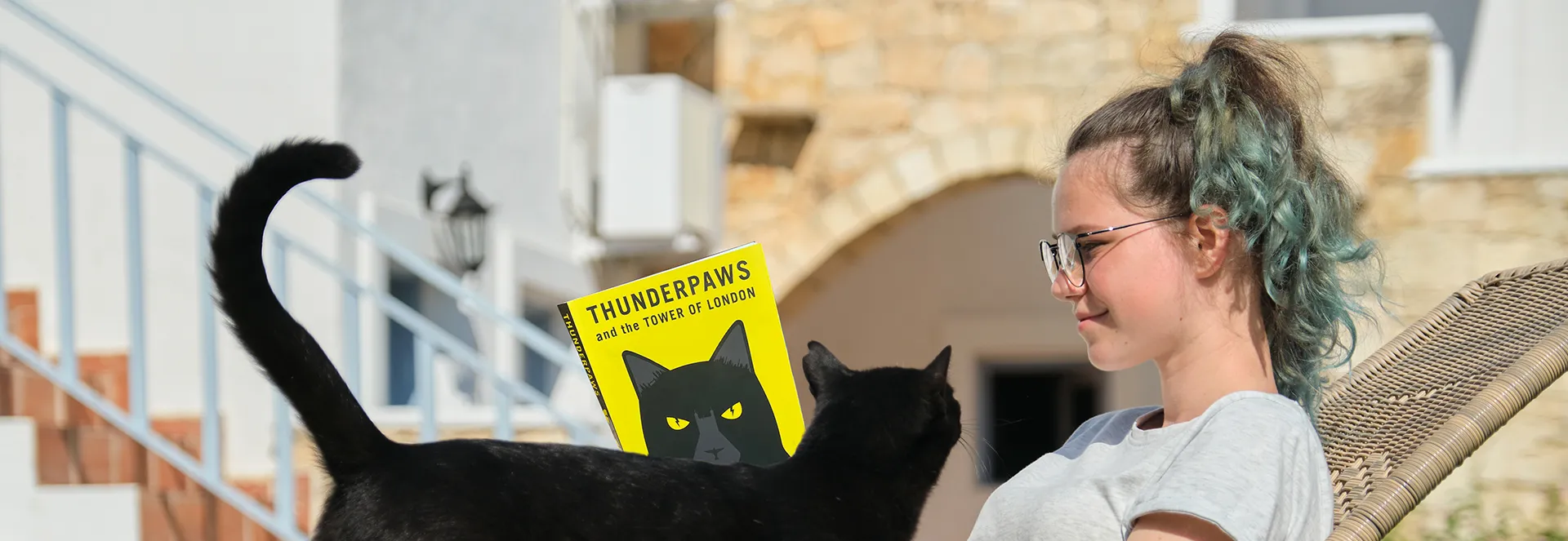 Person with glasses and a black cat, holding a book titled 'Thunderpaws and the Tower of London'.