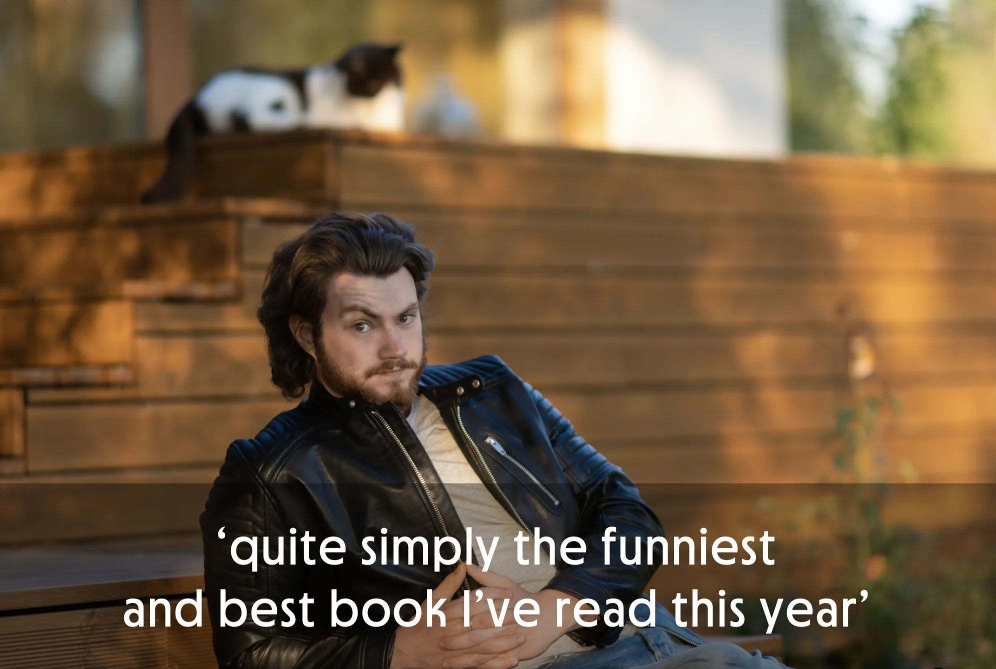 Man in leather jacket sitting outdoors with a cat in the background.