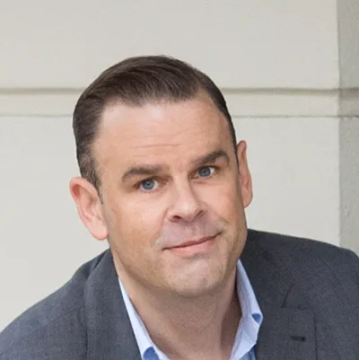 A man in a suit with short hair, smiling slightly.