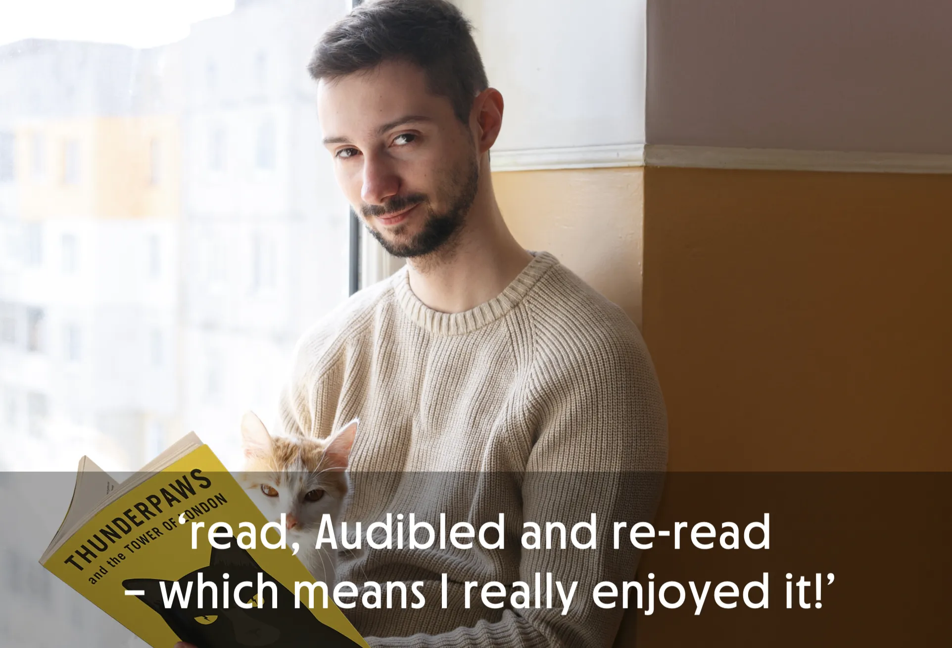 Man sitting by window holding a book with a cat peeking over it.