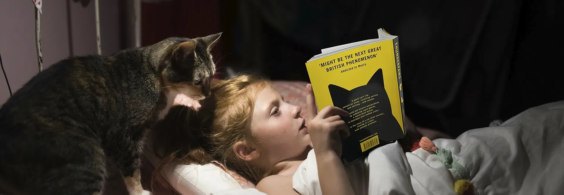 Child reading a book in bed with a cat nearby.