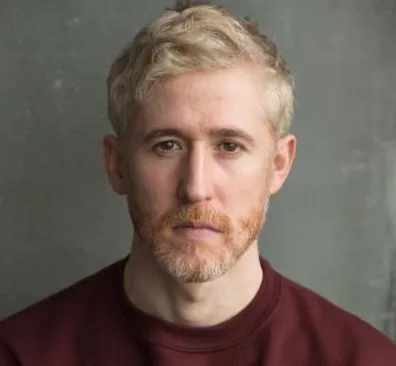 A person with short blond hair and a beard, wearing a maroon shirt, against a gray background.