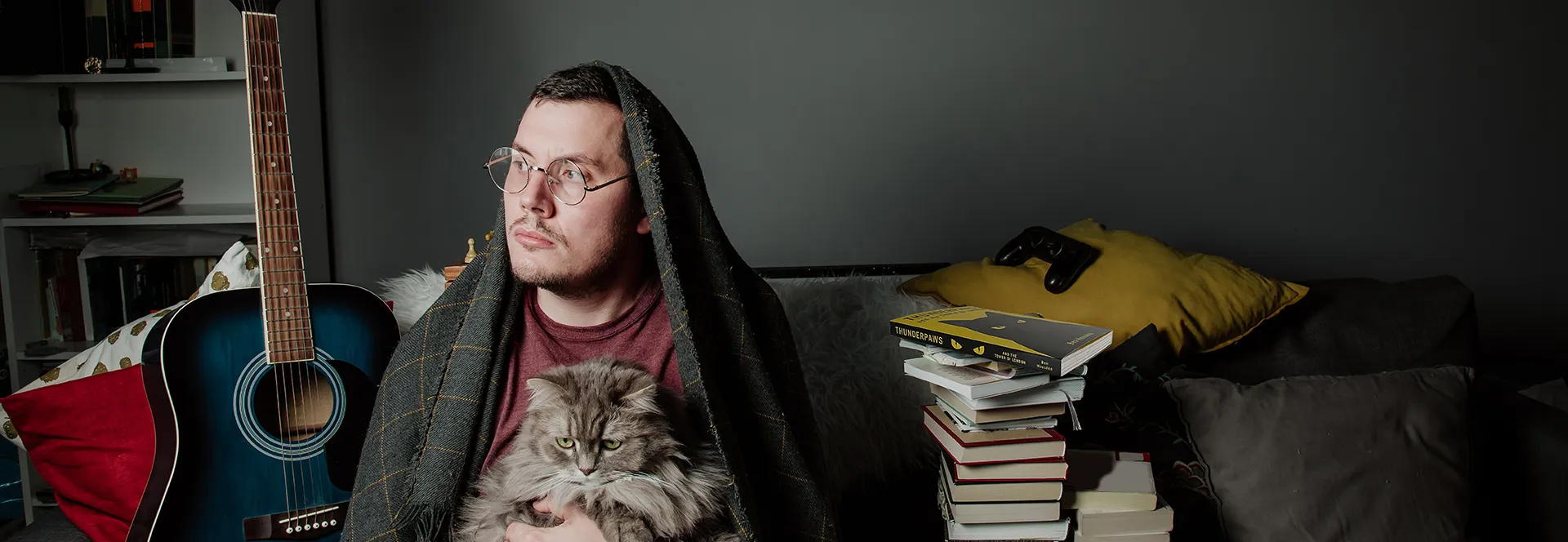 Person with glasses holding a cat, sitting by a guitar and stack of books.