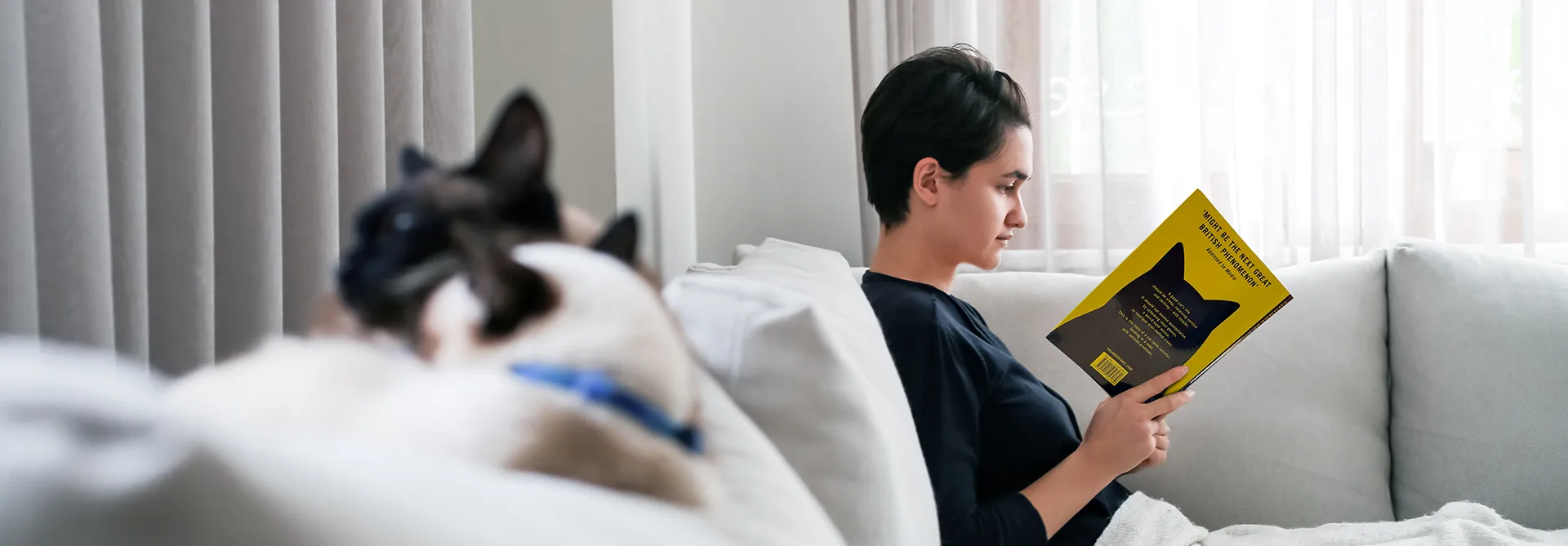 Person reading a book on a couch with a cat in the foreground.