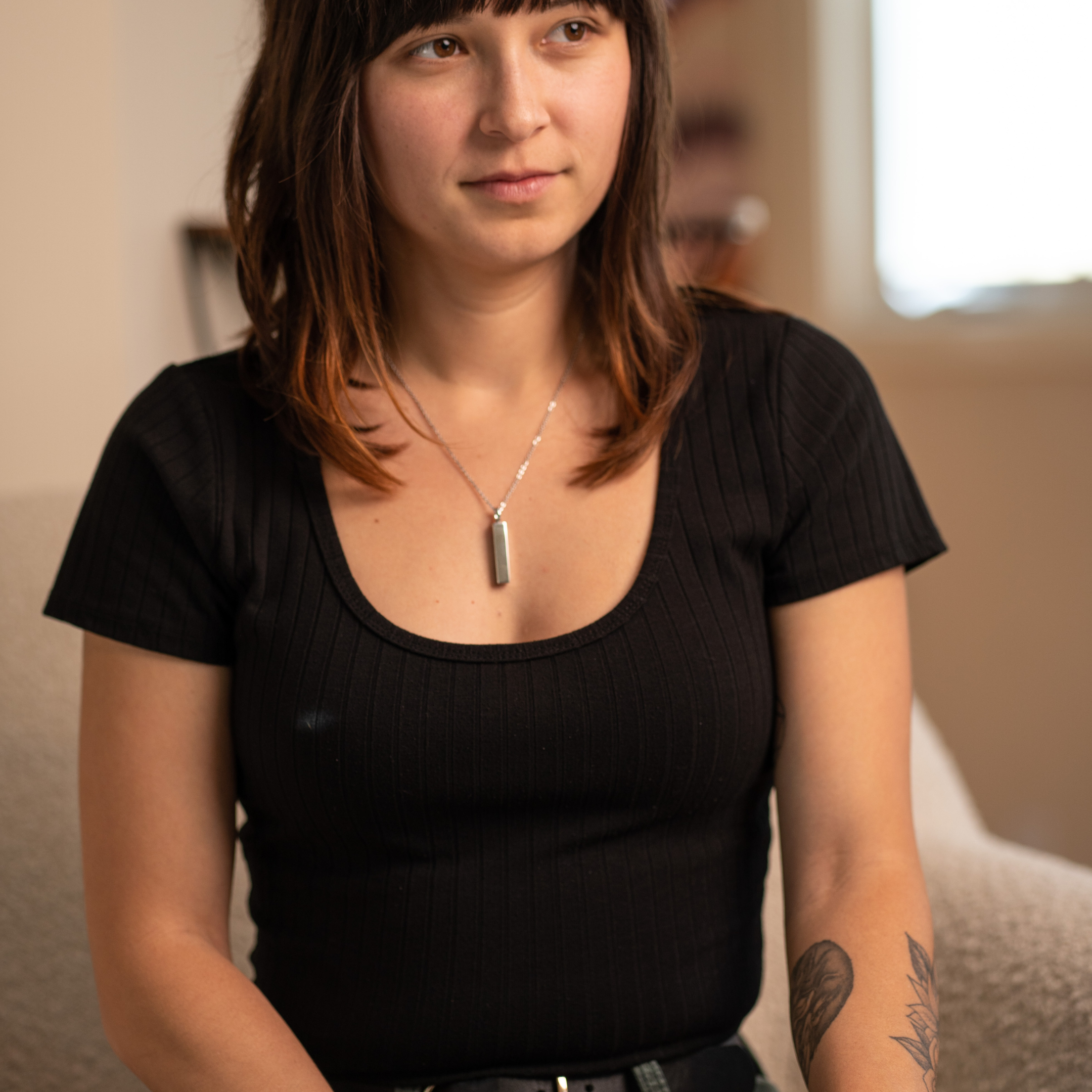 Woman with brown hair and a tattoo, wearing a black shirt, sitting indoors.