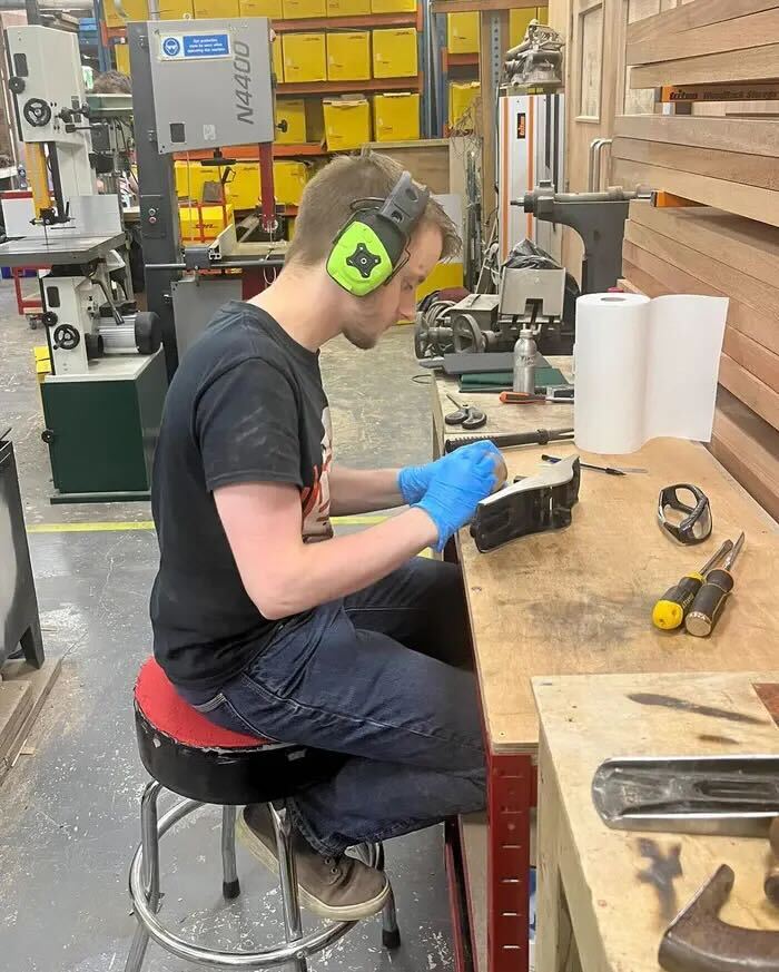 A man wearing ear protection and blue gloves works at a wooden workbench in a workshop.