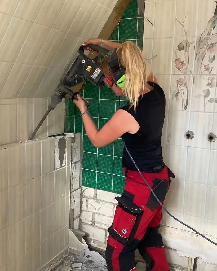 A woman in safety gear uses a demolition hammer to break tiles off a bathroom wall.
