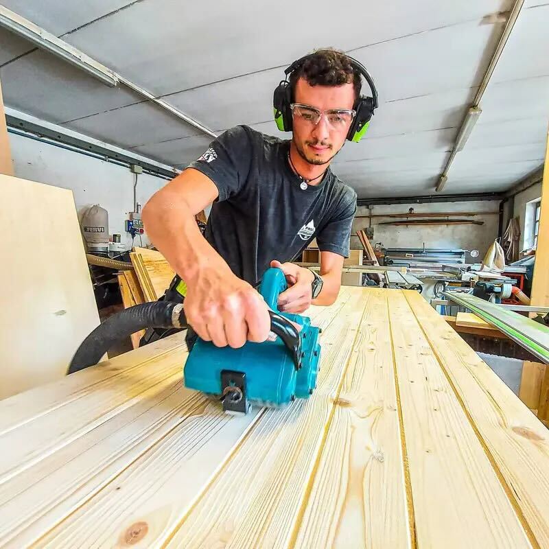 A man in a workshop wearing safety gear uses a power planer on a wooden surface.