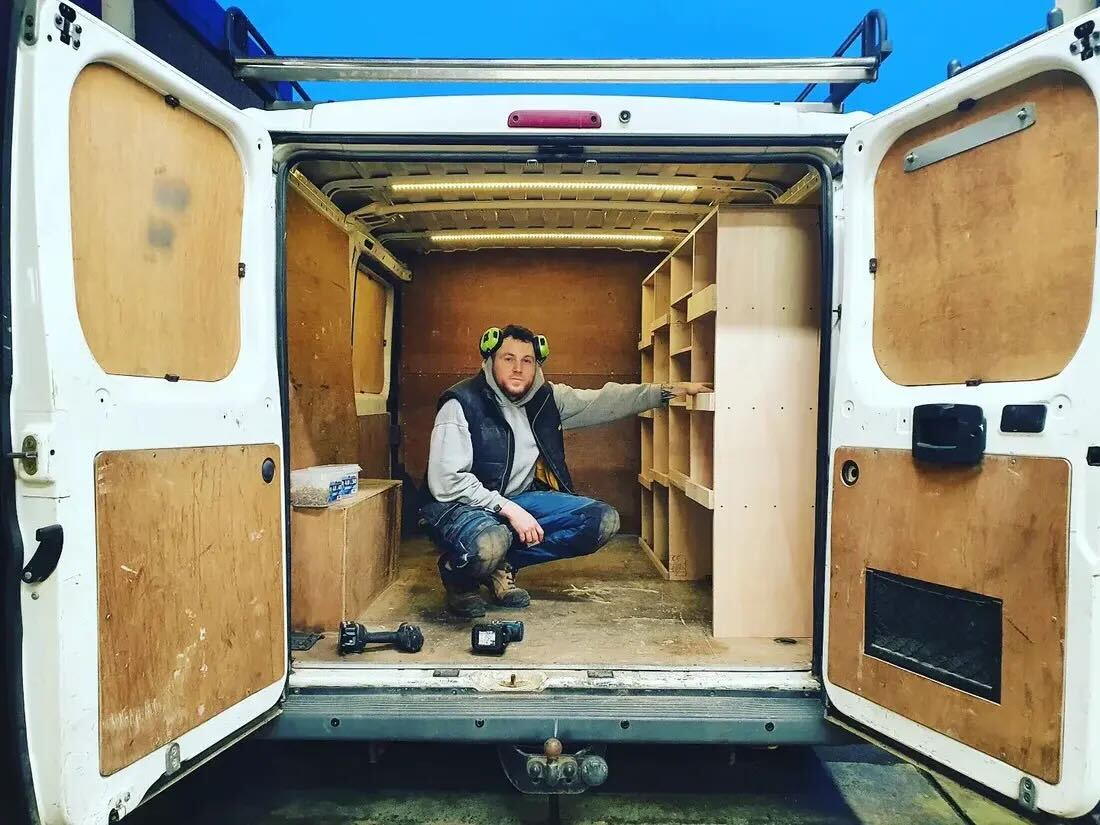 A man wearing ear protection installs a new wooden shelving unit in the back of a van.