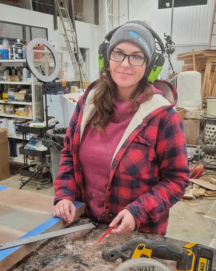 A woman wearing a beanie, glasses, and ear protection smiles while standing at a workbench in a workshop.