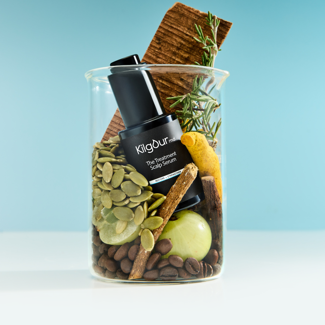 A beaker with scalp serum, seeds, herbs, and spices against a blue background.