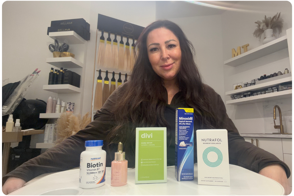 A woman sits at a table displaying several hair care and growth products in a salon setting.