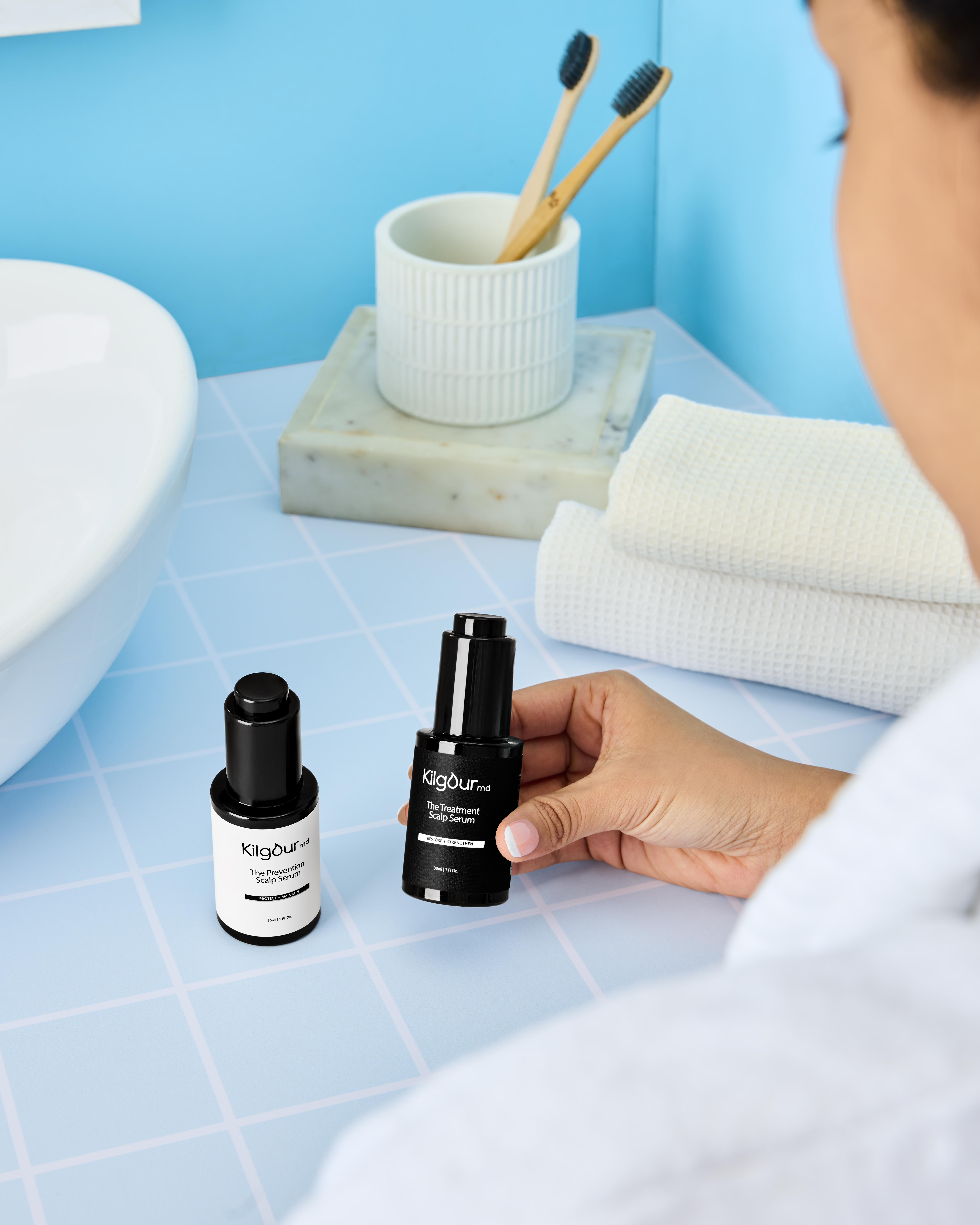 A person in a white robe holds a black bottle of scalp serum in a bathroom.