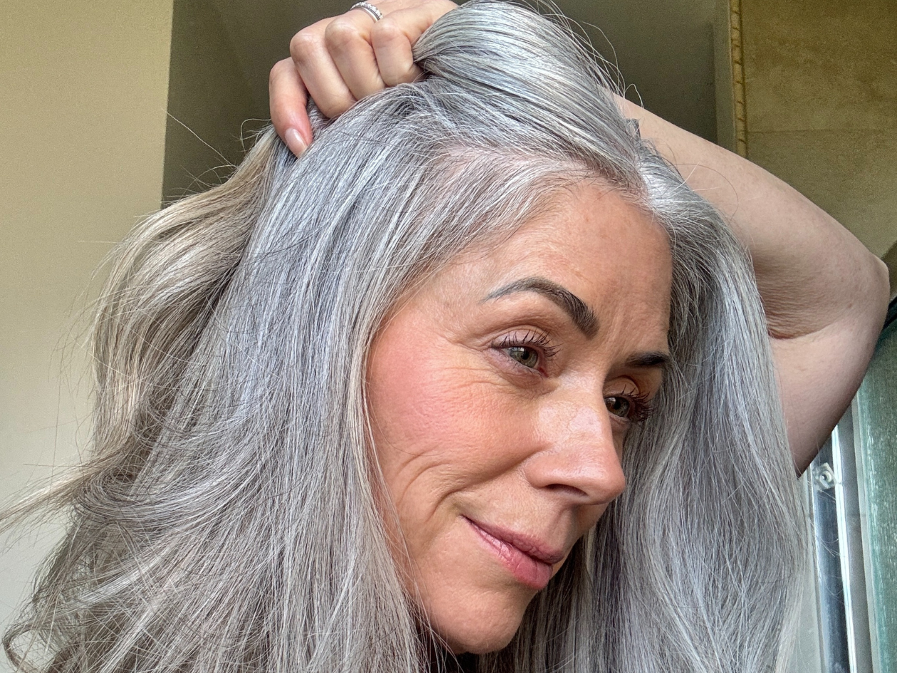 Person with long gray hair holding it back, smiling softly.