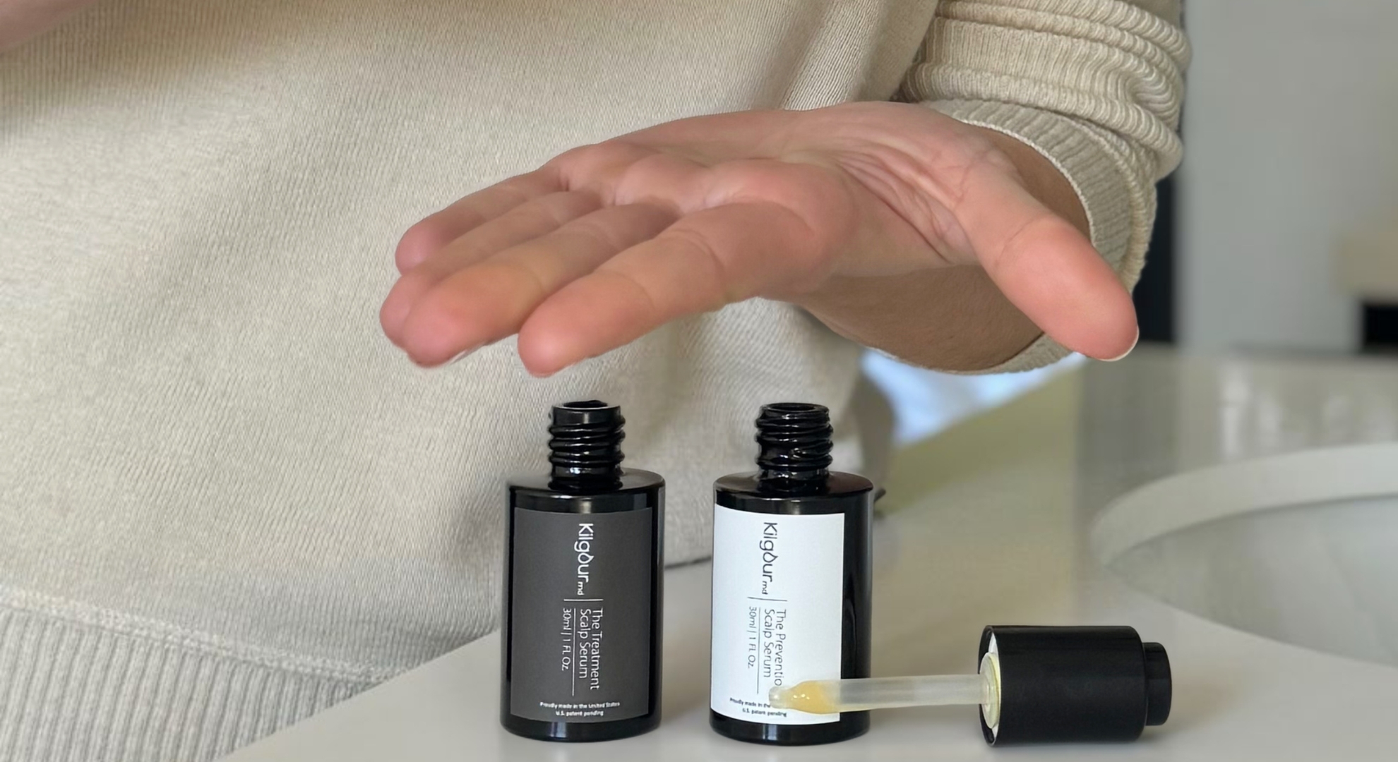 Hand holding a dropper above two black serum bottles on a counter.