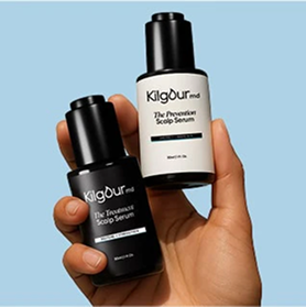 A hand holds two bottles of Kilgour MD scalp serum, one black and one white, against a blue background.