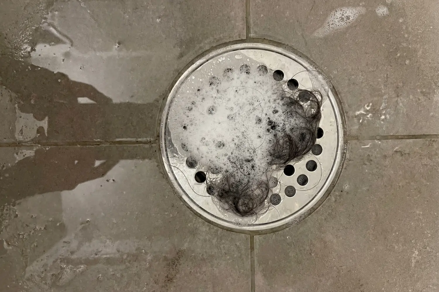 Clump of shed hair and foamy shampoo suds caught in a round shower drain on tiled floor