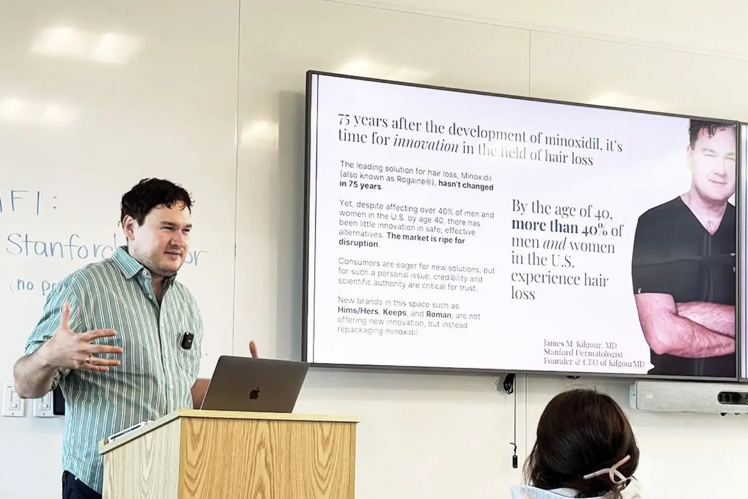 A man in a striped shirt gives a presentation about hair loss in a classroom setting.