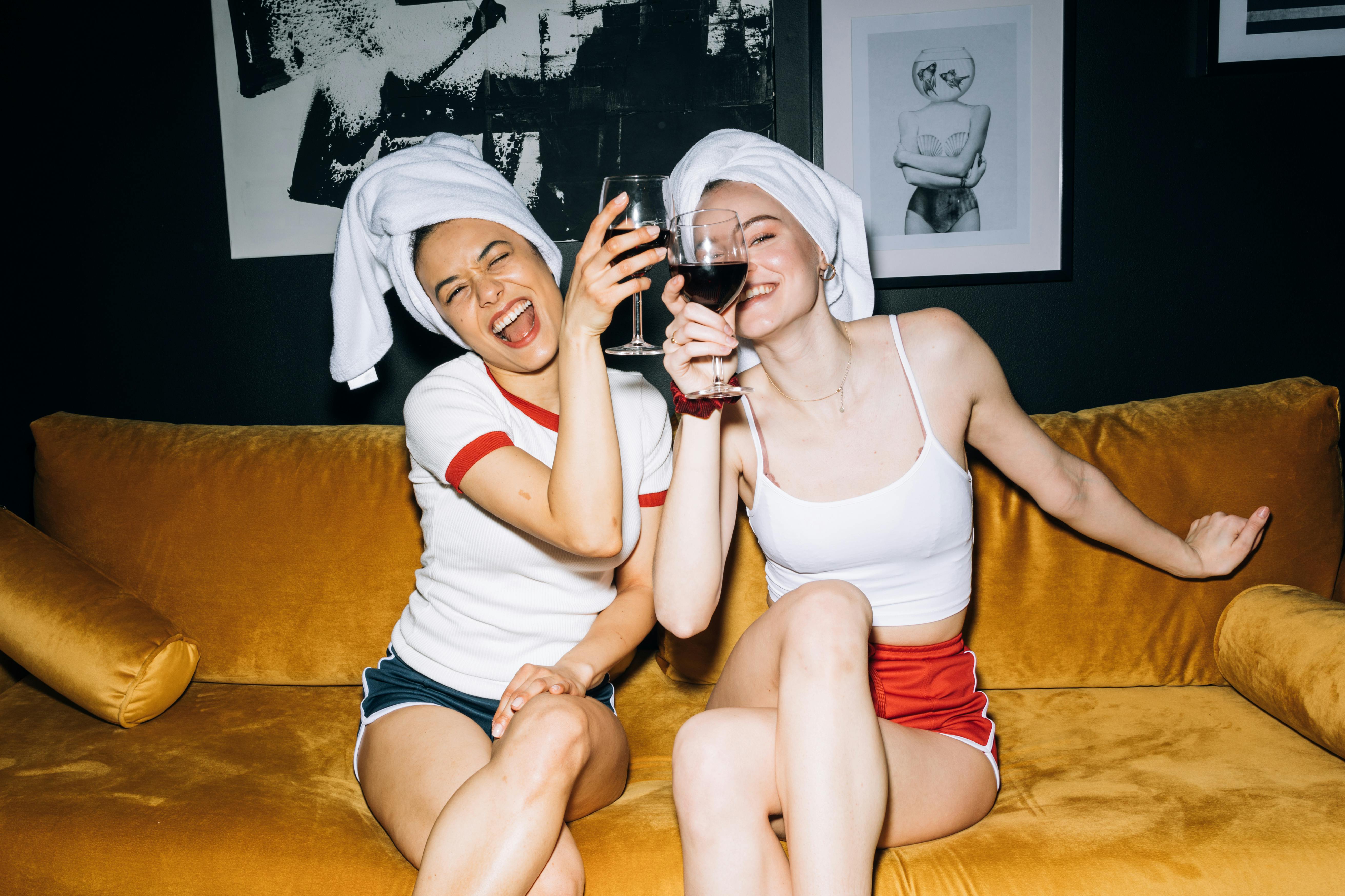 Two people with towels on their heads, laughing and holding wine glasses on a yellow couch.