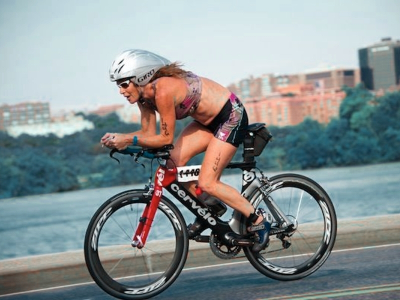 Cyclist in aerodynamic gear riding a Cervélo bike on a road with cityscape background.