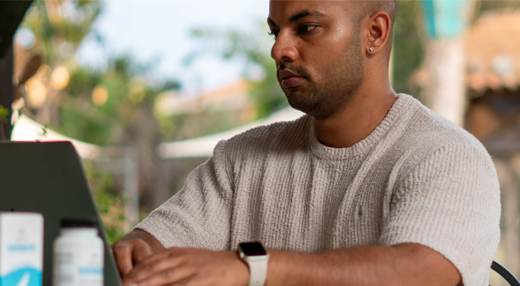 Man working on a laptop outdoors, wearing a smartwatch and casual sweater.