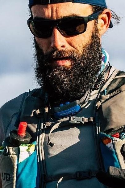 Man with a beard wearing sunglasses and outdoor gear, including a hydration pack.