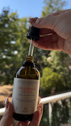 Hand holding a dropper above a brown bottle outdoors.