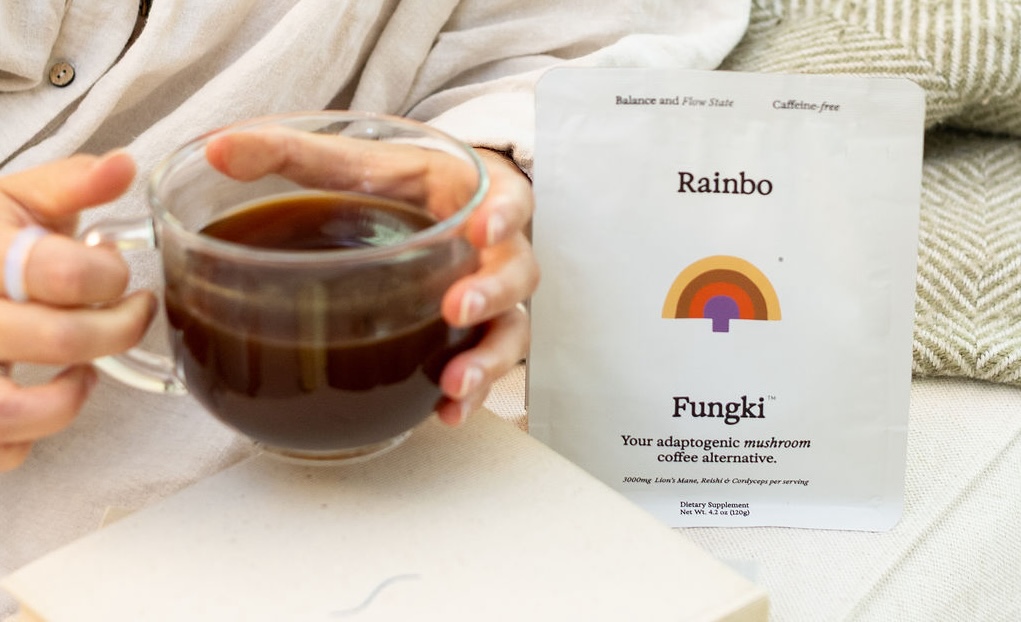 Person holding a glass cup of dark liquid next to a Rainbo Fungki product packet.