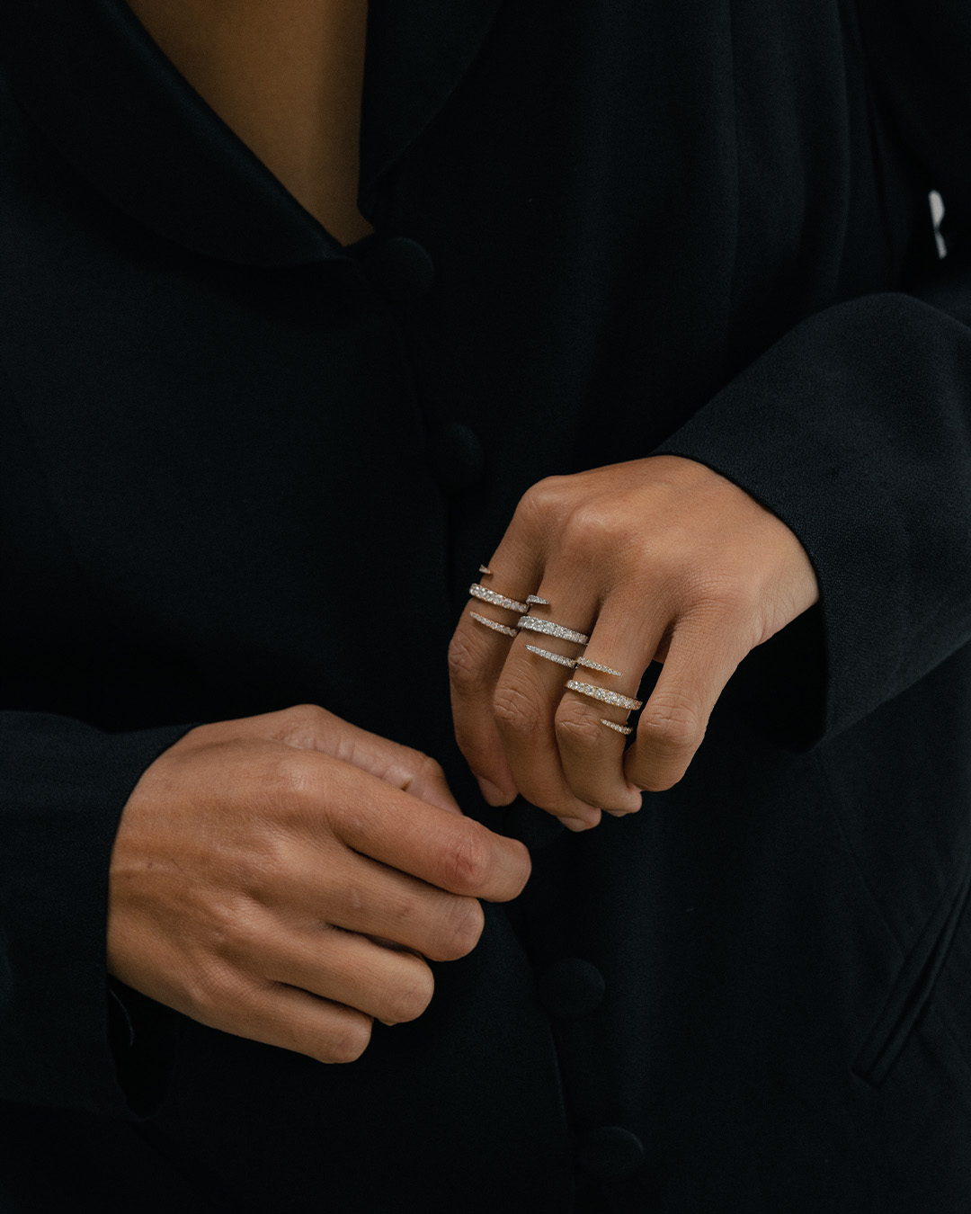 A close-up of a person's hands wearing a black jacket and several pointed, diamond-encrusted rings.