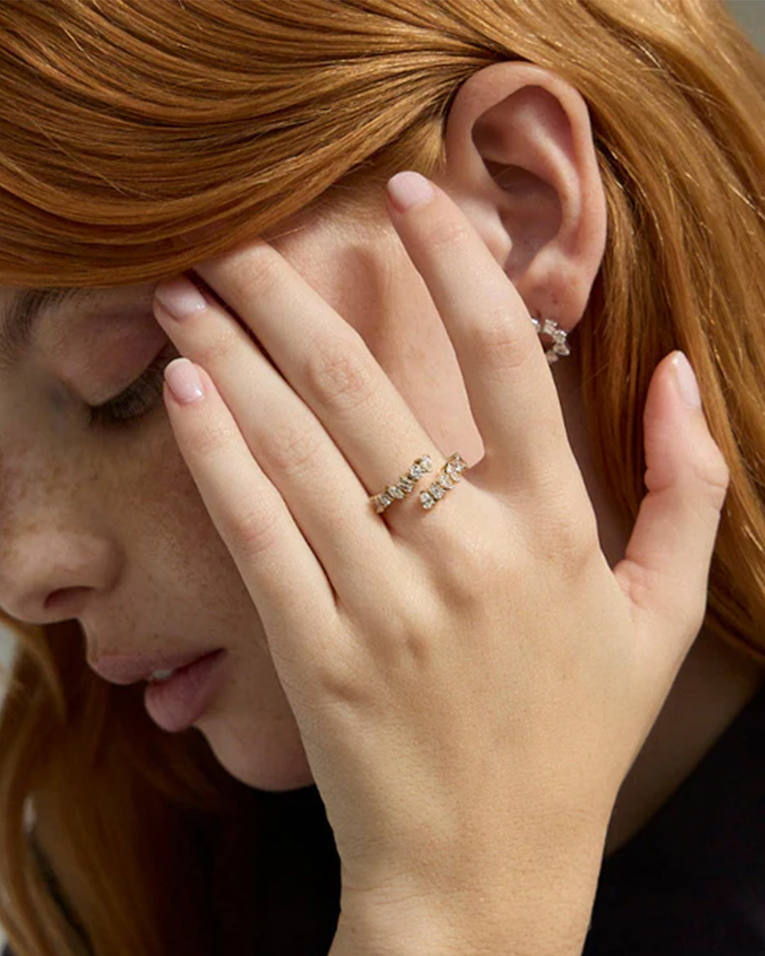 A close-up of a woman with red hair wearing a diamond ring and earring, with her hand to her face.