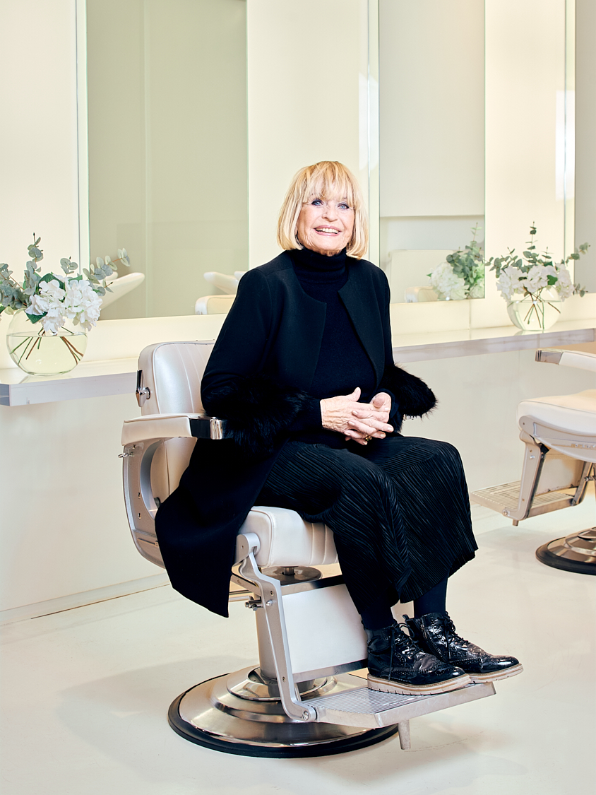 Smiling person seated in a salon chair, surrounded by mirrors and floral arrangements.