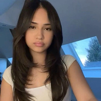 Person with long dark hair in a white shirt, sitting indoors near a window.