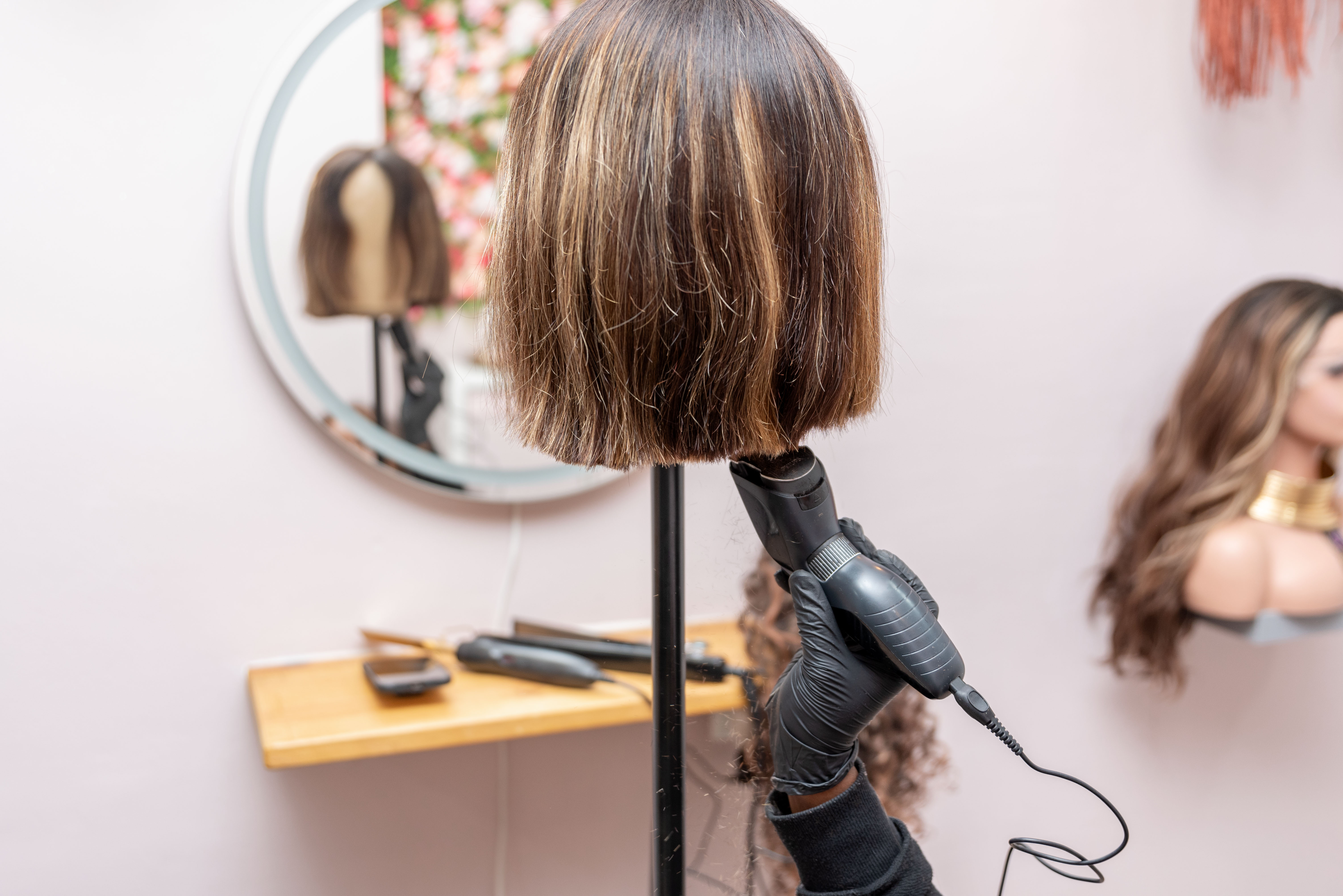 Gloved hand styling short brown wig with a curling iron, seen in front of a mirror.