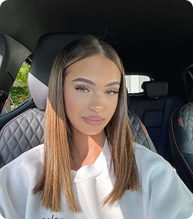Person with straight brown hair sitting in a car, wearing a white top.