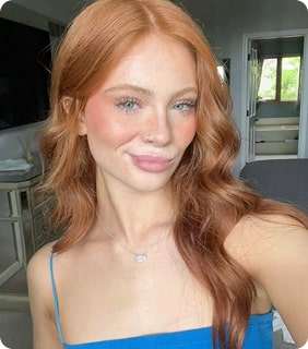 Person with long red hair smiling indoors, wearing a blue top.