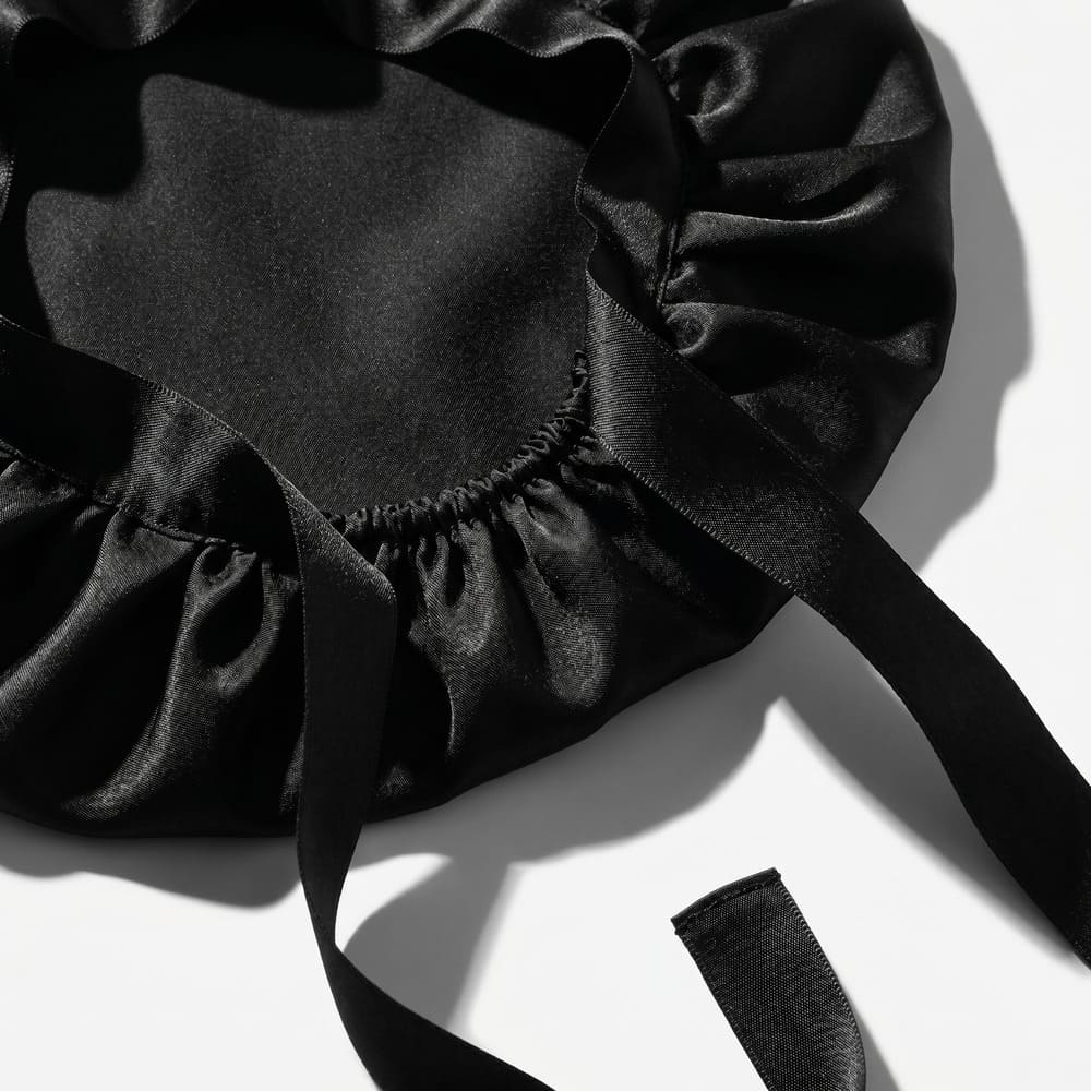 A close-up of a black satin bonnet with a gathered edge and ribbon ties on a white surface.