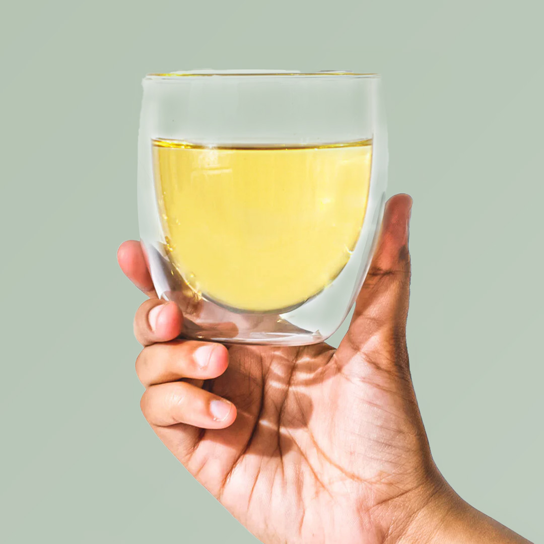 Hand holding a glass of yellow liquid against a light green background.