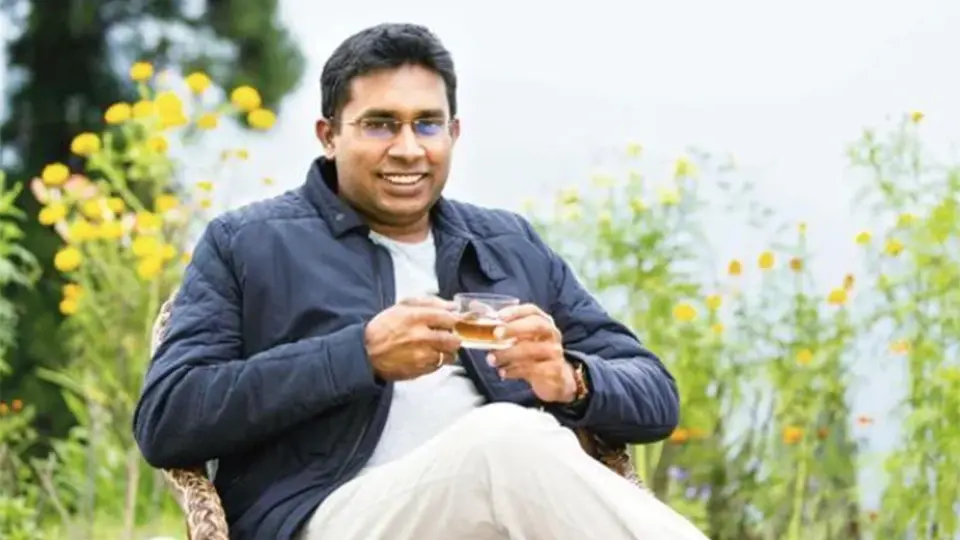 A smiling man in glasses sits in a chair outdoors, holding a small glass of tea.