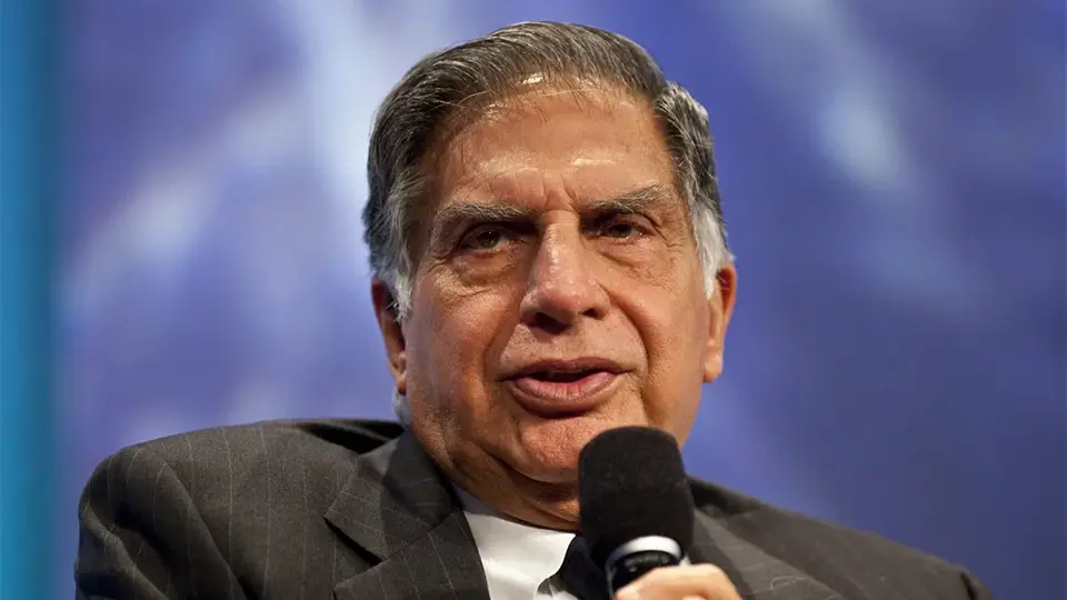 A headshot of an older man in a suit speaking into a microphone against a blue background.
