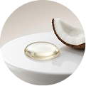 A single drop of oil sits on a white surface with a piece of fresh coconut behind it.