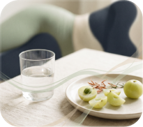 A glass of water on a table next to a plate of whole and sliced green amla fruit.
