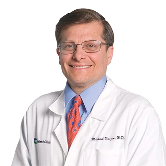 Person in a white lab coat with glasses and a blue shirt, smiling.