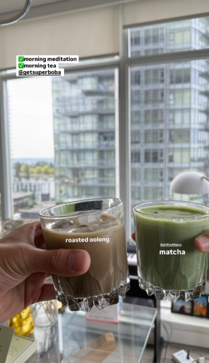 Two hands holding glasses of roasted oolong and matcha tea in front of a city view.