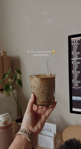 Hand holding an iced coffee in a cozy room with a plant and computer screen.